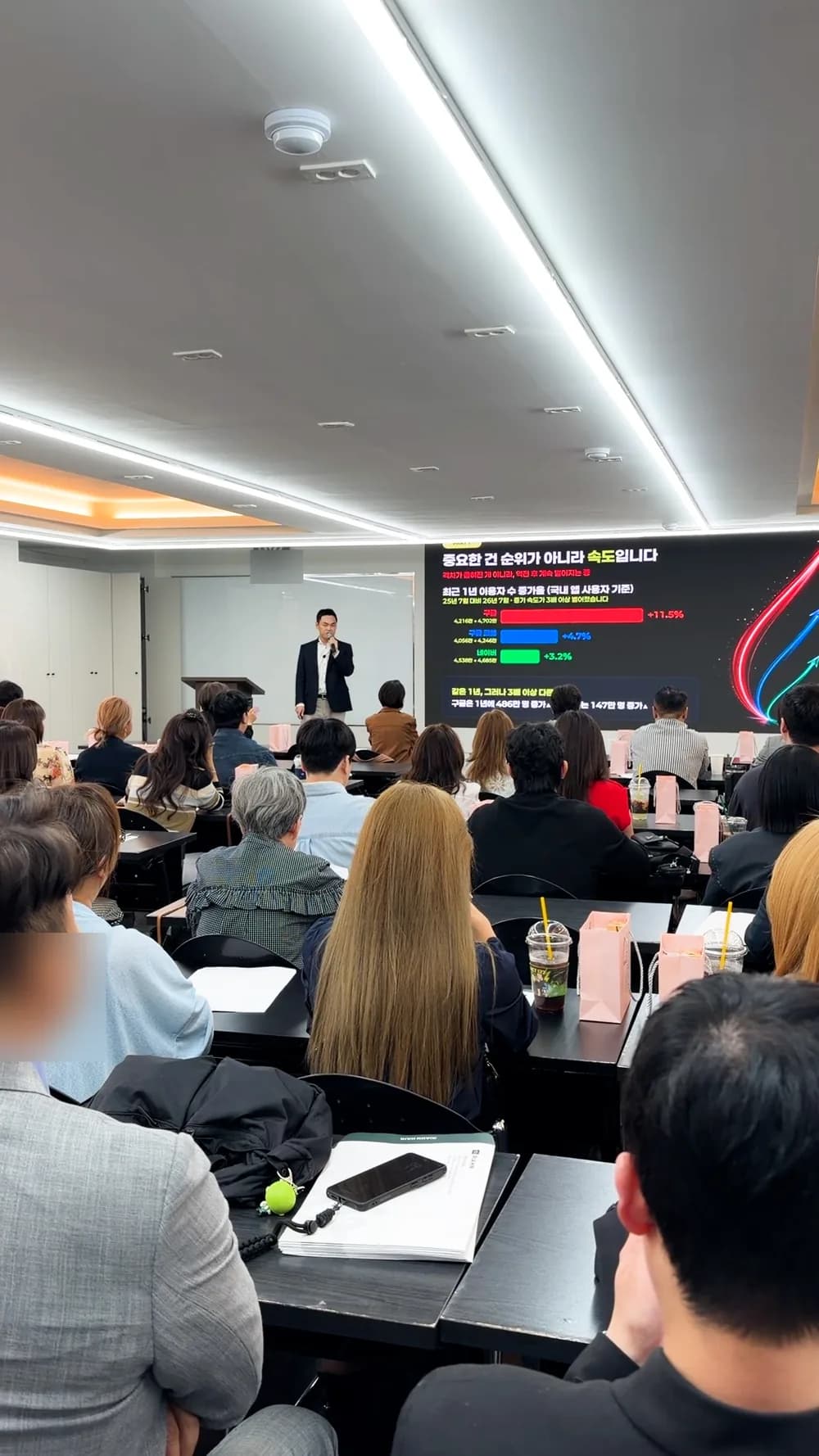 강연장 뒤쪽에서 본 전경. 참석자들이 자리를 채우고 있고 앞쪽 화면에 이용자 수 비교 슬라이드가 떠 있다